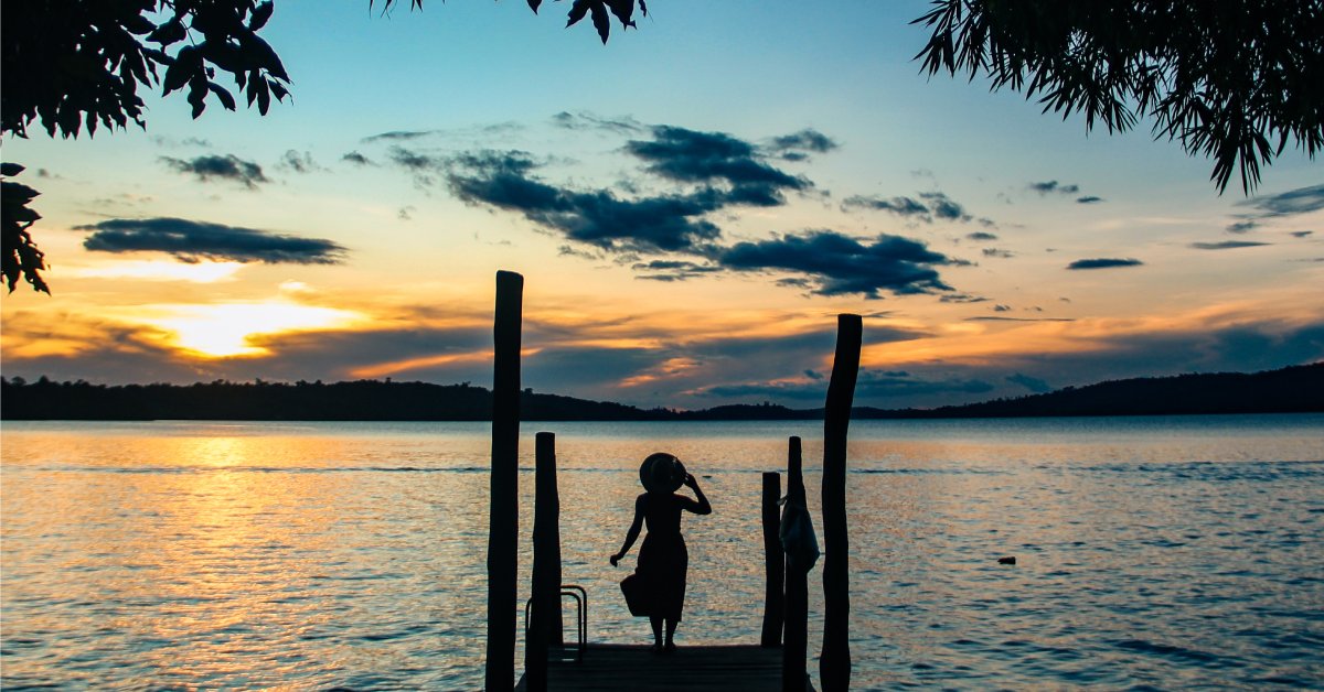 The silhouette of a woman in a dress and wide-brimmed hat standing on a dock and looking out at the sunset.