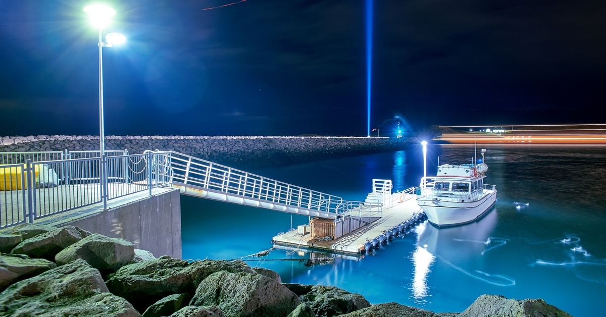A boat floating next to a small dock at nighttime. The dock has a long, slim walkway. Bright lights illuminate the area.