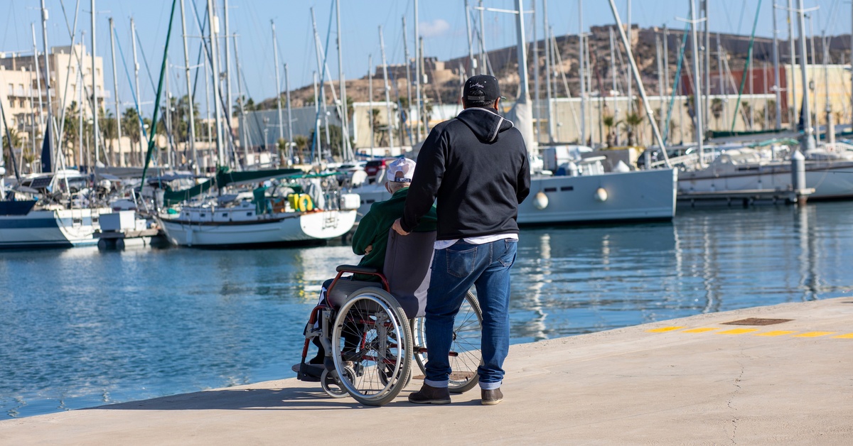 A person in a wheelchair sitting at the edge of a marina on a sunny day. Another person is standing behind them.